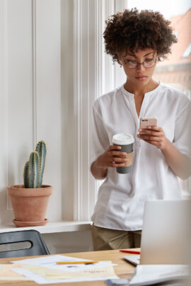 Serious attractive dark skinned girl copywriter works on advertising campaign, checks email via cell phone, stands near desk with documents and laptop computer, connected to wireless internet